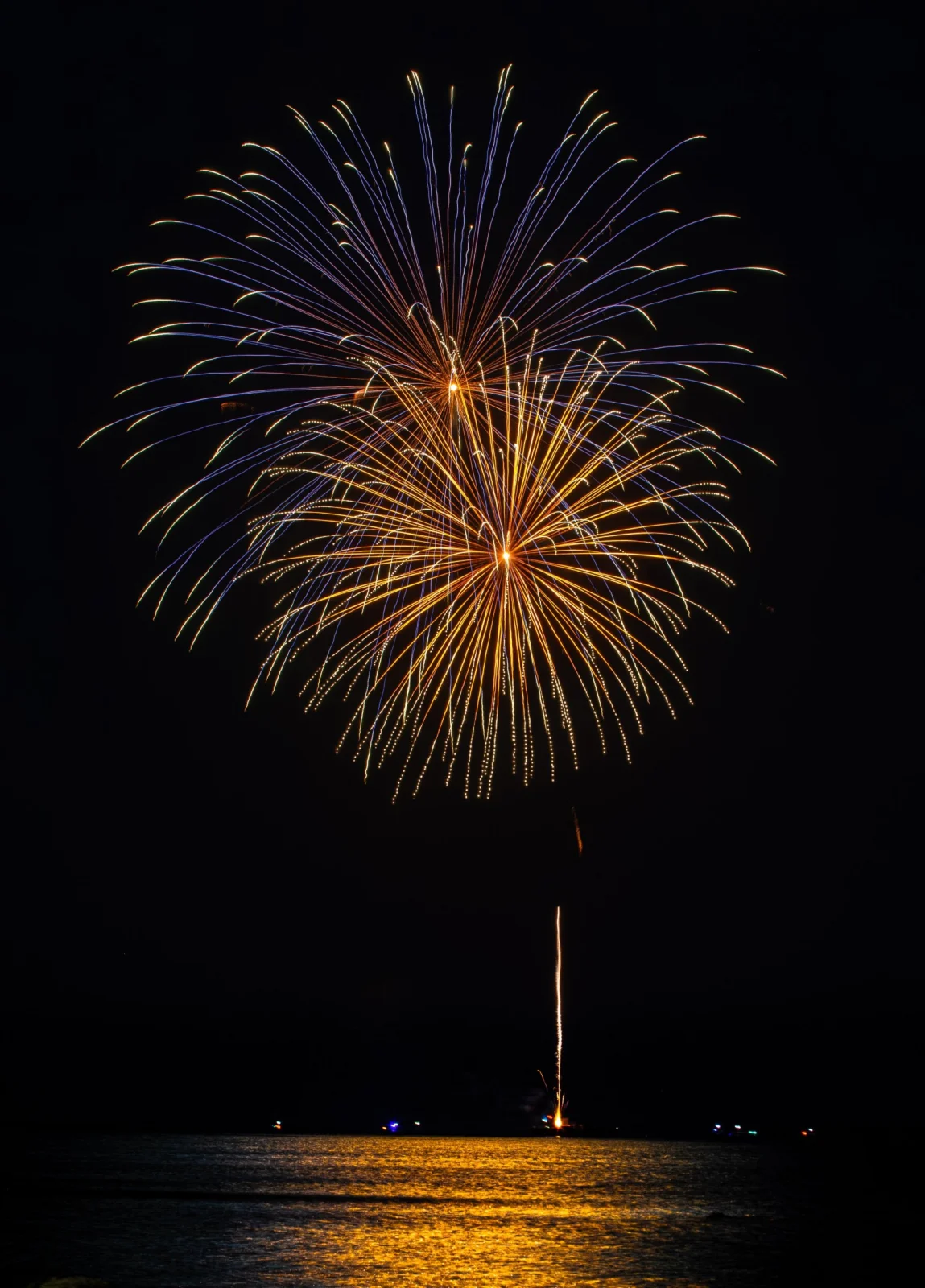 Gold and violet fireworks bursting over dark water, with a new shell rising from a boat
