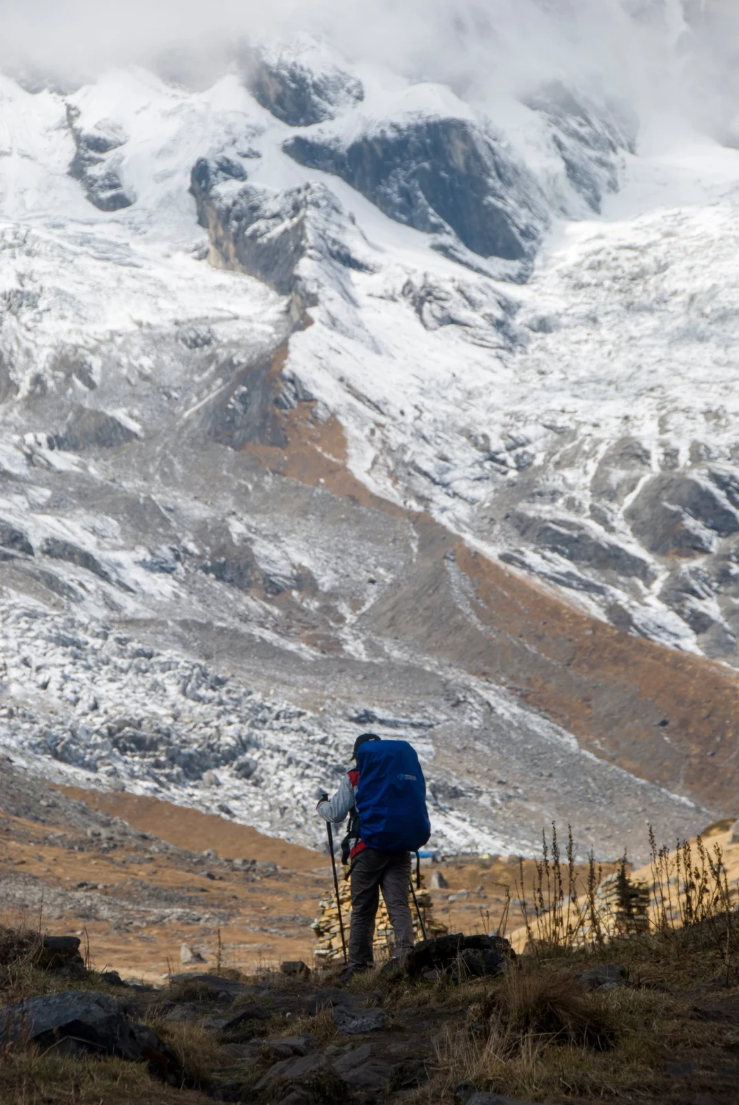 A hiker with a blue pack and trekking poles walking toward a snow-covered mountain face